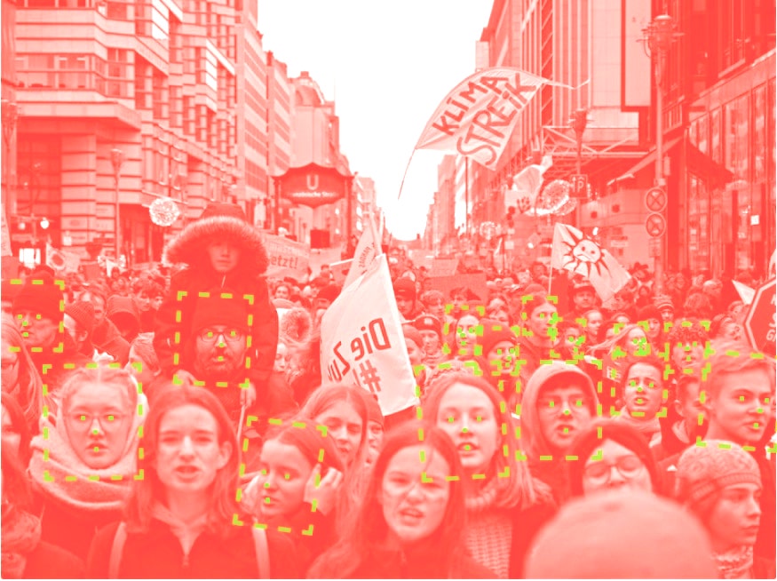 A monochrome image of a crowd of people in a Berlin street, holding placards and banners. Facial recognition bounding boxes surround a number of the faces, with dots identifying the location of the eyes and mouth.